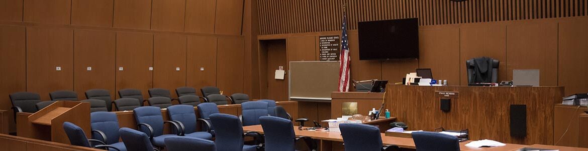 Picture of an empy court room. Empty chairs surround a large table in the front of the court. All Empty chairs face forward infront of the Judge's seat.
