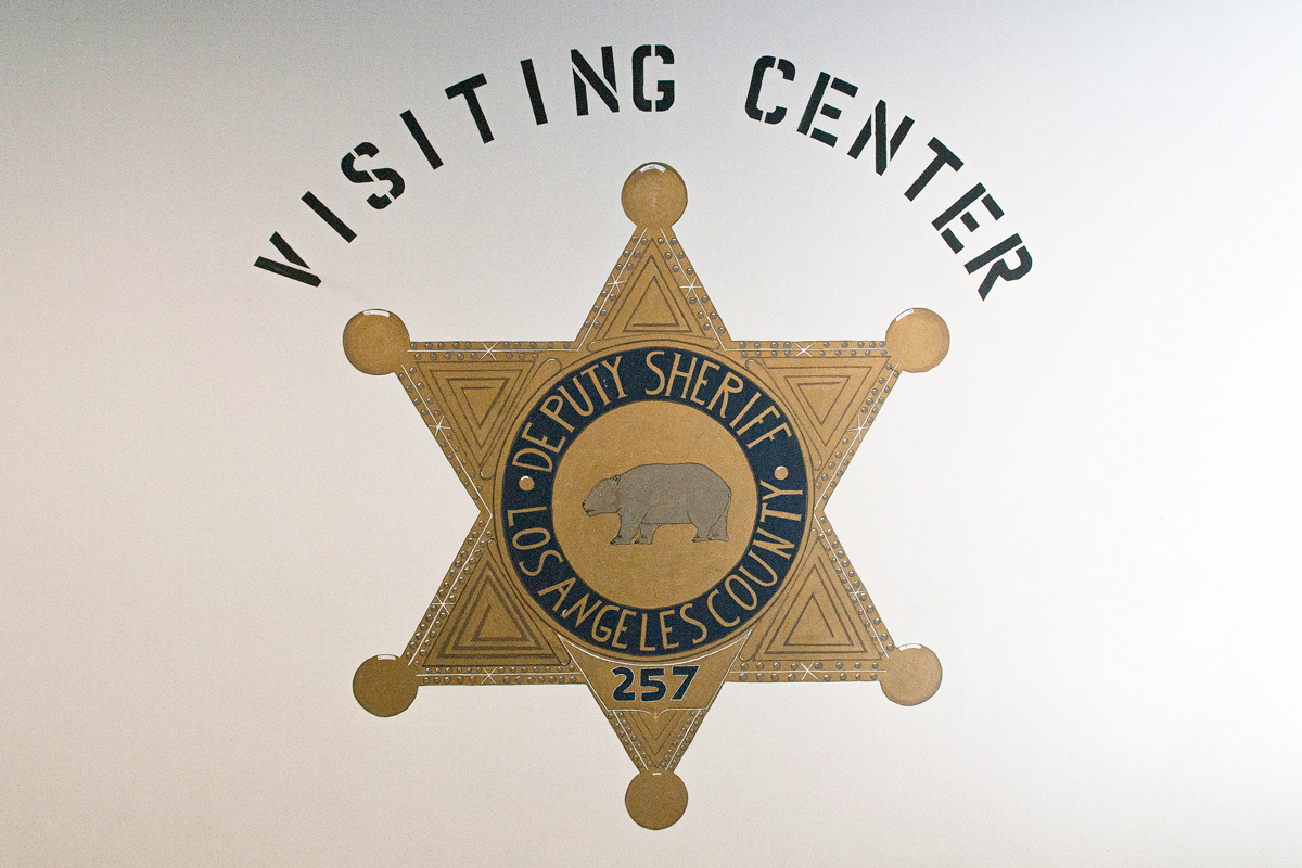 Picture of hand painted sheriff's badge, painted on a white wall in the C R D F Jail. The words above read, "Visiting Center".