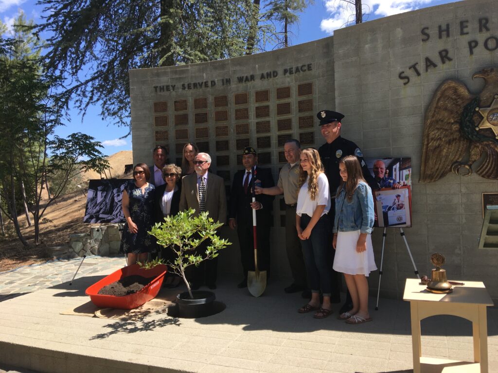 Family standing in front of memoral wall, small tree is being planted, Sheriff Alex Villanueva is holding a shoval.
