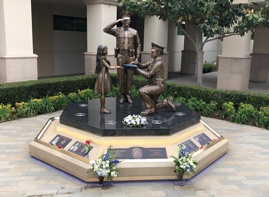Image of the Buena Park Police departent memorial of fallen officers. Bronze statue of a saluting officer, another officer on one knee presenting a folded flag to a little girl. all cast in bronze.