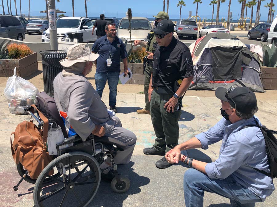 MET team deputy helping a homelss man in a wheelchair. there are three people from Vetran affairs talking to a man in a large white sun hat, he is dressed in a long sleeve gray shirt,the wheel chari has an orange backpack on the back, the homeless man is in his 60s.