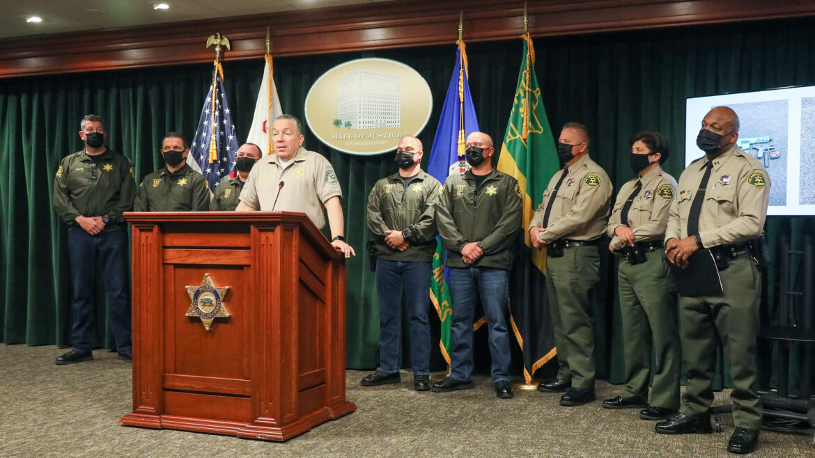 picture of Sheriff Alex Villanueva standing at a podium speaking to the audience. The podium is brown and has the Sheriff's Badge on the front. There are 8 other personel from O S S behind the Sheriff. A screen is off to the right behind O S S Captain with pictures of two guns on the screen.