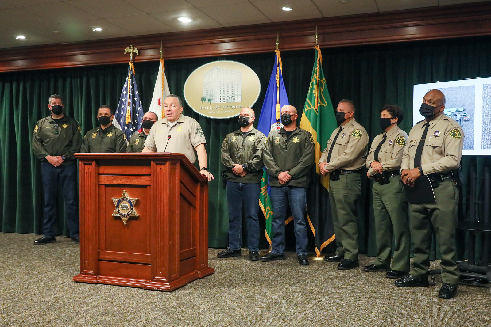 picture of Sheriff Alex Villanueva standing at a podium speaking to the audience. The podium is brown and has the Sheriff's Badge on the front. There are 8 other personel from O S S behind the Sheriff. A screen is off to the right behind O S S Captain with pictures of two guns on the screen.
