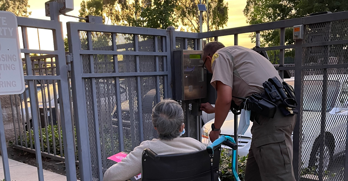Image of a woman in a wheel chair being helped into a gate by a Deputy. The deputy is looking at a panel on the security gate trying to buzz the person into the complex.