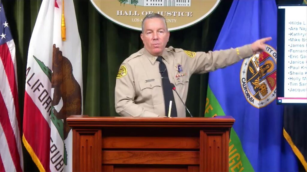 Sheriff Alex Villanueva is standing behind a wooden Podium and is pointing off to the right to a screen.