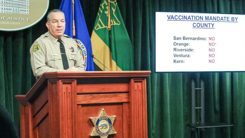 Sheriff Alex Villanueva is standing behind a wood Podium speaking to media. He is standing infront of a green curtain next to County and California flags. There is a TV screen, on the screen the slide reads: "Vaccination Mandates by County: San Bernardino, no; Orange, no; Riverside, no; Ventura, no; Kern, no;'