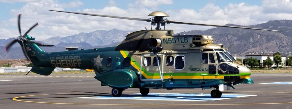 Image of Search and rescue heliocoptor on a runway. The heliocoptor is green and gold, is very large with a slideing door on the side where Search and Rescue personnel operate a wench. The markings on the heliocoptor are the Sheriff's star, and the words "Rescue 5".