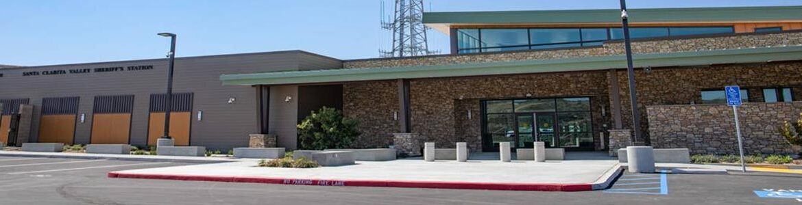 Image of the front parking lot and entrance of Santa Clarita Sheriff's station. The building has a long flat roof that is painted green on the trim. the entrance is of Beige brick and has a large sliding glass door.