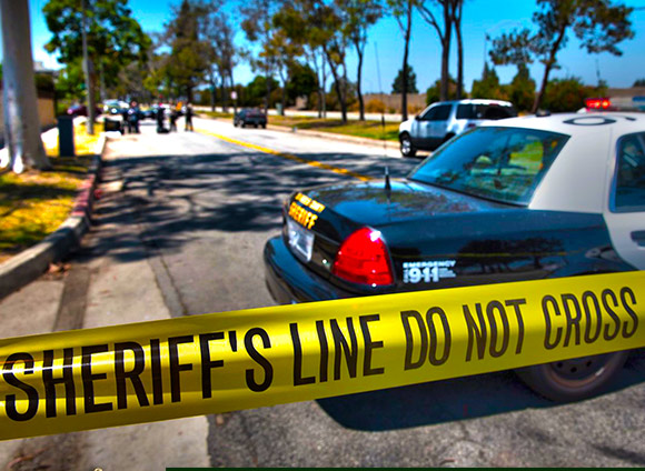 Image of a taped off street. Yellow tape reads: Sheriff's line do not cross. end of a police car is in the front and in the distance a congregation of sheriff's Deputies.