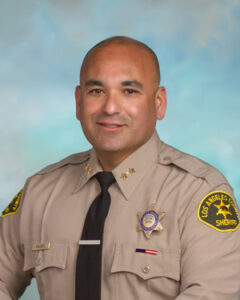 Portrait of Chief valdez posed from the waist up. In front of a blue background. He is dressed in he Sheriff's uniform, tan, with a black tie.