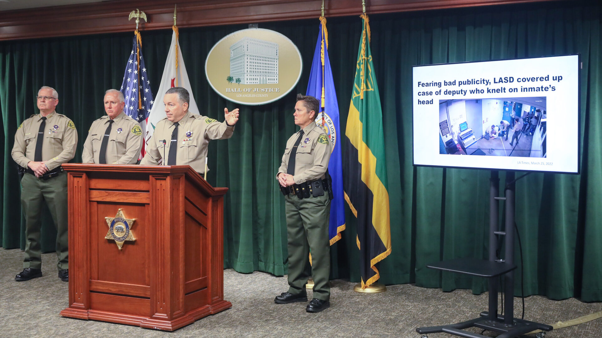 Sheriff Alex Villanueva is in a tan and green uniform standing infront of a wooden podium with the Sheriff's badge attached to the front. he is pointing to a screen on his right. He has two of his assistant Sheriff's on his left and one assistant Serhiff on his right.