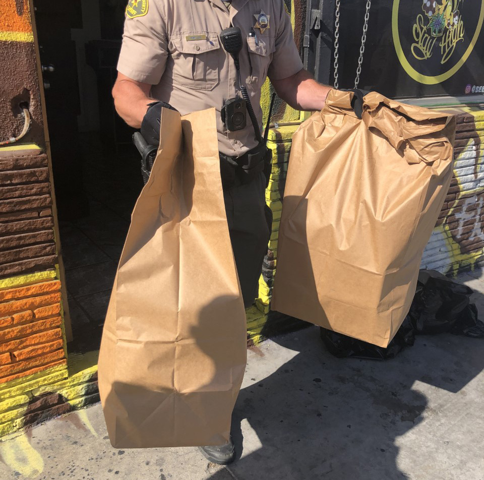 deputy is walking out of a business door holding two paper bags full of confiscated drugs.