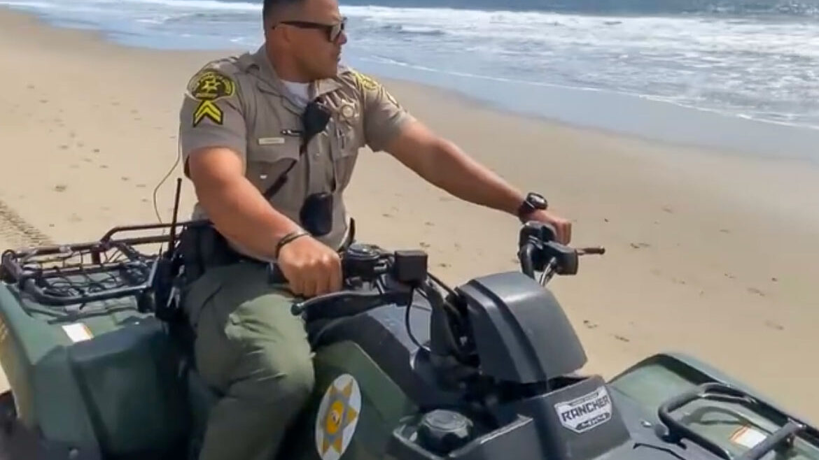 Image of a deputy wearing a tan Sheriff's Uniform riding a 4 wheel motorcycle. he is riding on a beach with the ocean and skyline behind him.
