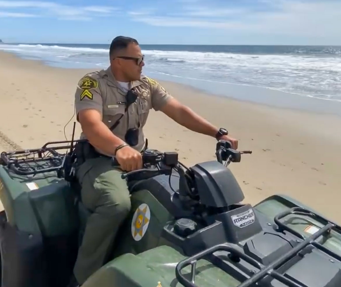 Image of a deputy wearing a tan Sheriff's Uniform riding a 4 wheel motorcycle. he is riding on a beach with the ocean and skyline behind him.