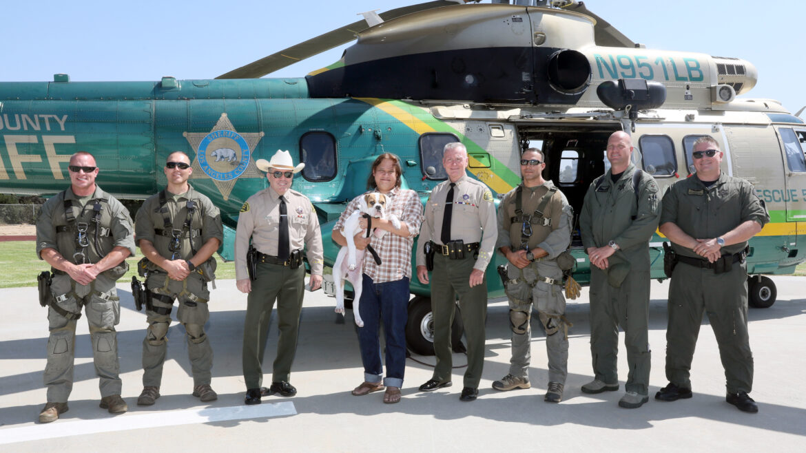Air 5 Heliocoptor crew stands with Sheriff Villanueva and the rescued hiker and his dog infornt of the large green and gold heliocoptor.