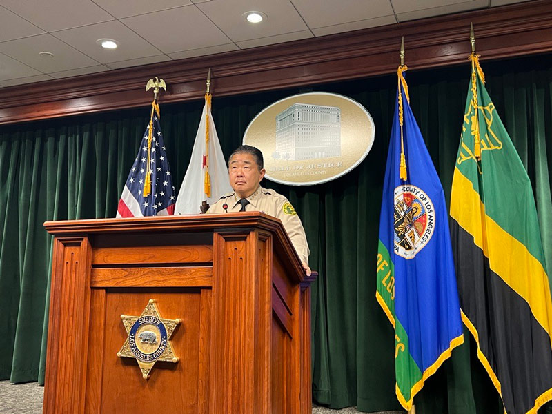 Image of Undersheriff Murakami standing at a wooden podium. The podium is in a conference room with a green backdrop.