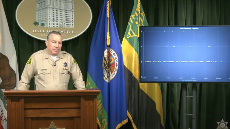 image of Sheriff Villanueva, standing behind a podium, a TV screen is off to his left shoulder showing a graph.