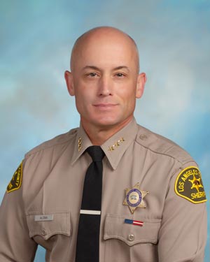 Assistant Sheriff Aloma is wearing a tan long sleeve shirt with a black tie, badge on left side of shirt, sitting in front of a blue background.