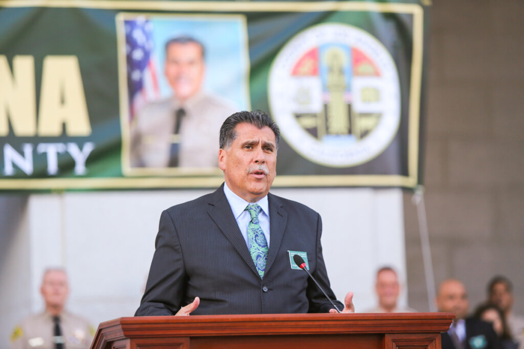 Picture of Sheriff Luna in a blue suit and green print tie with a white shirt. standing infront of a podium in front of the hall of justice. A large banner of Luna is in the background hanging off of the columns of the building