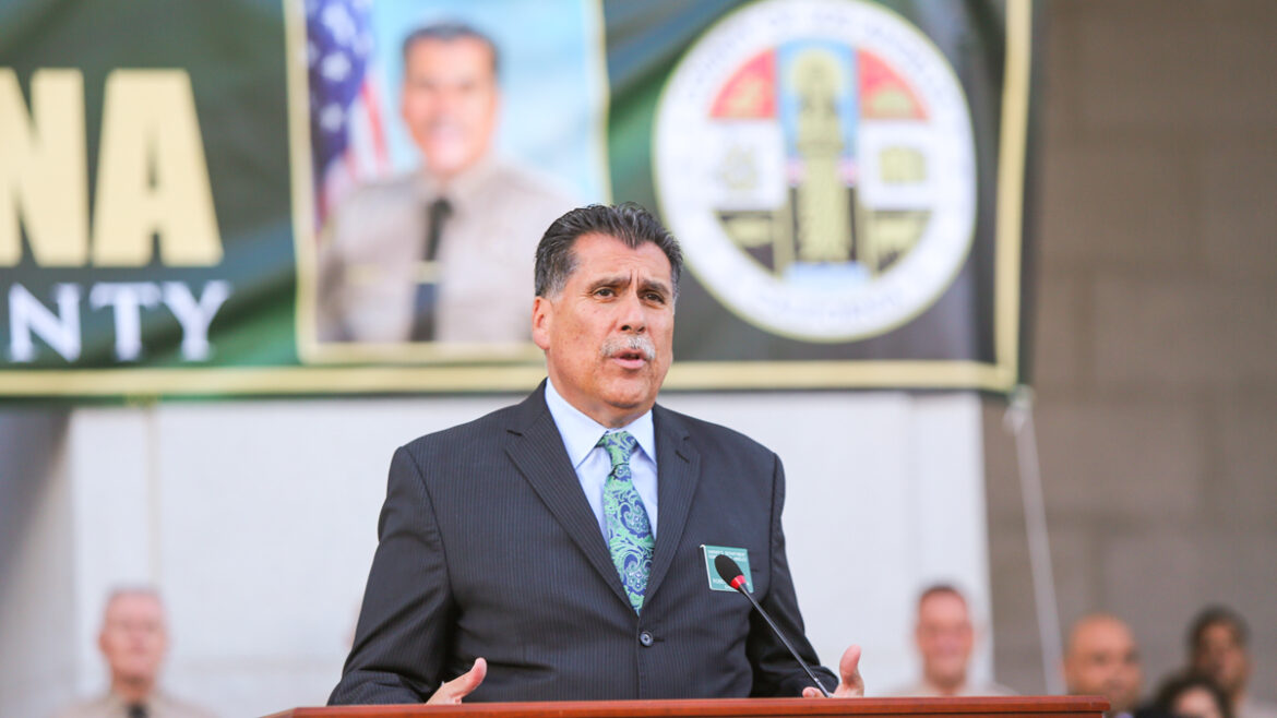 Picture of Sheriff Luna in a blue suit and green print tie with a white shirt. standing infront of a podium in front of the hall of justice. A large banner of Luna is in the background hanging off of the columns of the building