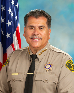 Sheriff Luna is wearing a tan long sleeve shirt with a black tie, badge on left side of shirt, sitting infront of an American flag, sitting in front of a blue background.