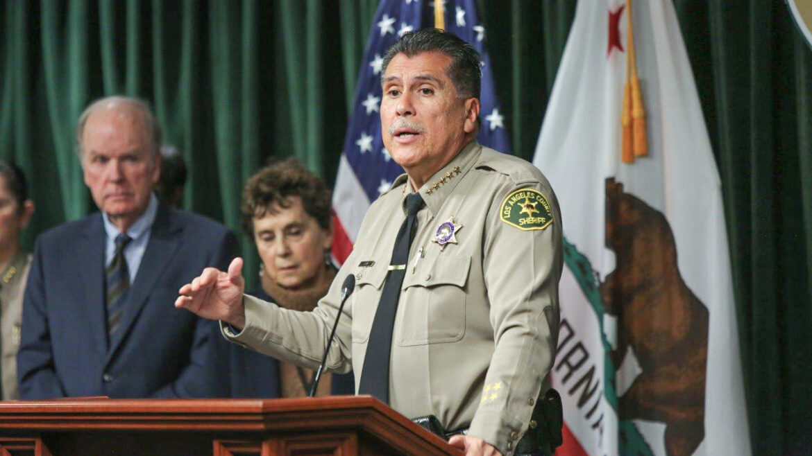 Sheriff Luna is speaking at a podium. He is dressed in a tan uniform, long sleeve tan buttondown shirt with a black tie. He is standing infront of the California state flag and the US flag.