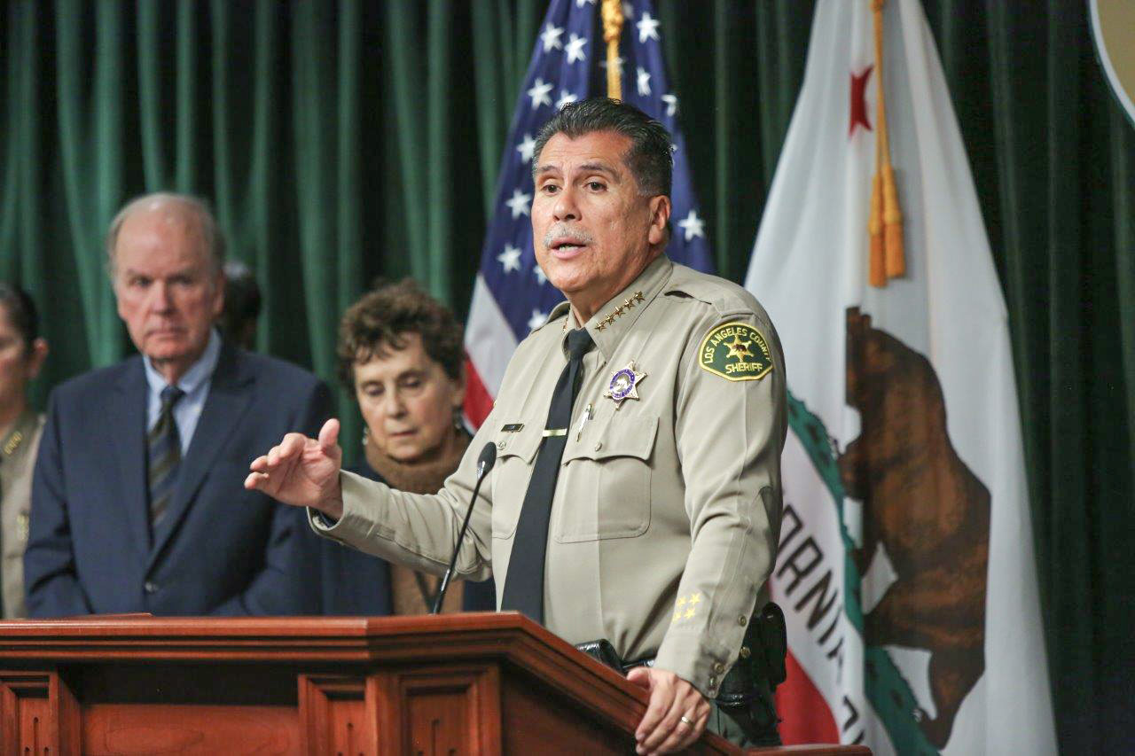 Sheriff Luna is speaking at a podium. He is dressed in a tan uniform, long sleeve tan buttondown shirt with a black tie. He is standing infront of the California state flag and the US flag.