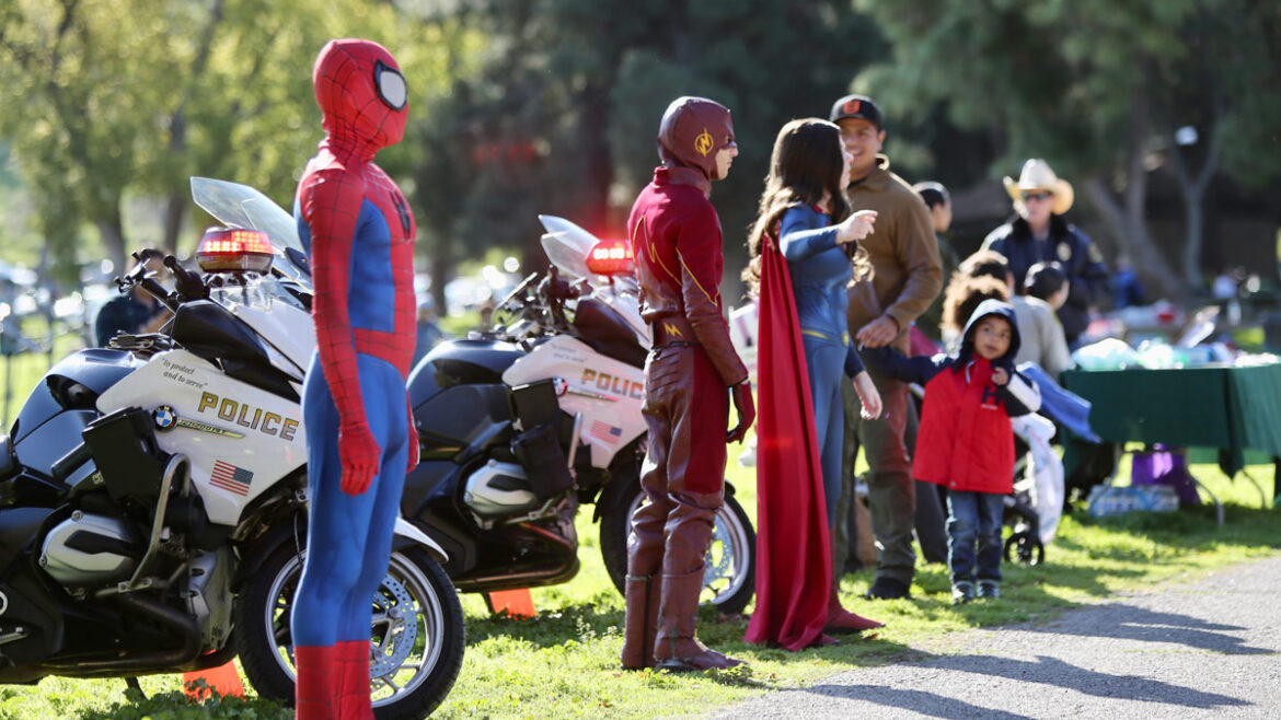 outdoor picture of Sheriff's motorcycles parked along a race path. People dresses as Spiderman, Flash and Supergirl are standing facing the racers.