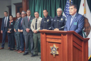Sheriff Robert luna in a blue suit standing behind a wooden podium. LASD staff as well as LAPD staff stand behind him.