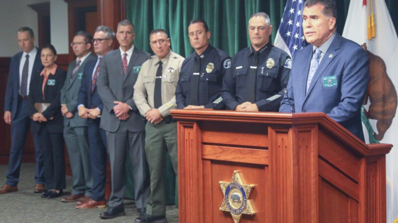Sheriff Robert luna in a blue suit standing behind a wooden podium. LASD staff as well as LAPD staff stand behind him.