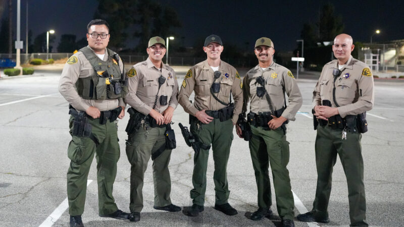 5 deputies in uniform posing shoulder to shoulder in a parking lot for a picture at night.