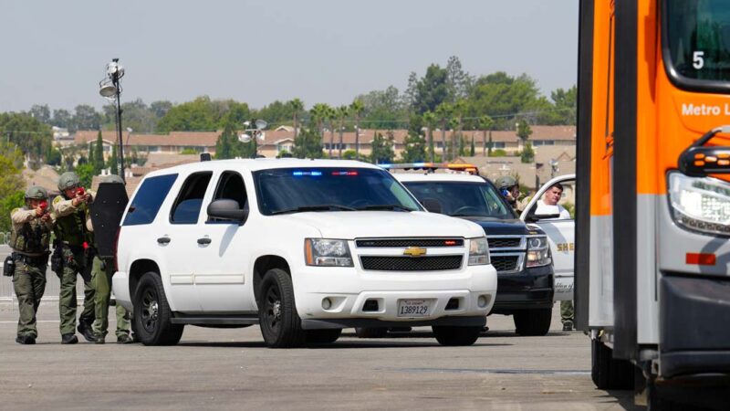 Three deputies in safety gear pointing training weapons from behind a white SUV at a bus parked in a parking lot training exercise.