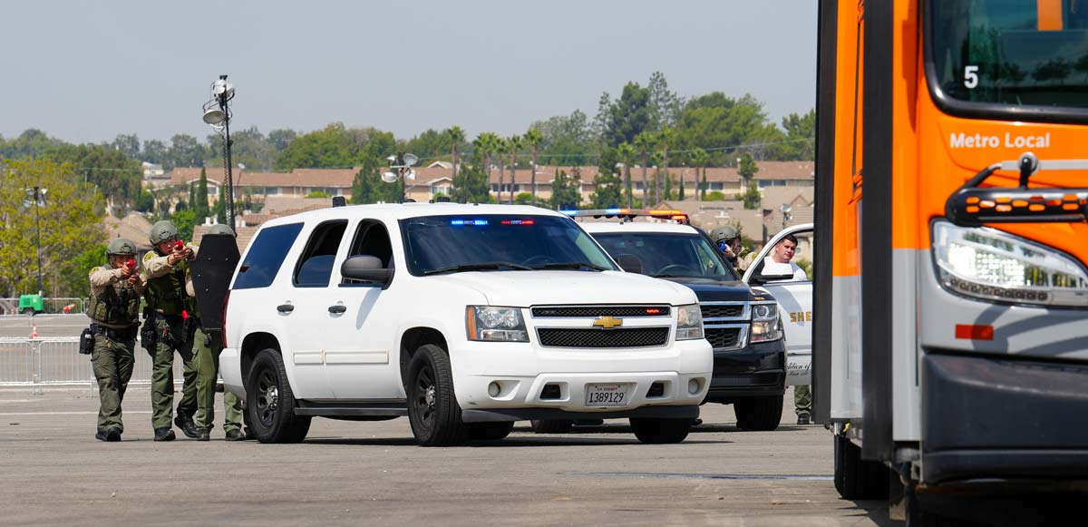Three deputies in safety gear pointing training weapons from behind a white SUV at a bus parked in a parking lot training exercise.