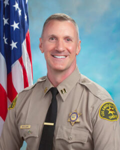 Captain Moulder is wearing a tan long sleeve shirt with a black tie, badge on left side of shirt, sitting in front of a blue background, the american flag is behind him, over his right shoulder.