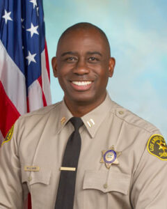 Captain Williams is wearing a tan long sleeve shirt with a black tie, badge on left side of shirt, sitting in front of a blue background. There is a hanging american flag in the background behind him on his right side.