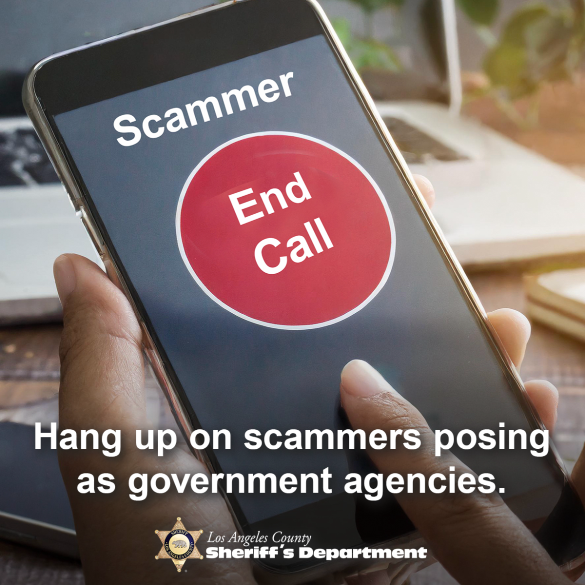 A hand holding a cell phone; on the screen, the caller ID says "Scammer" with a large red "End Call" button below
