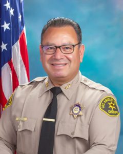 Chief Mendoza is wearing a tan long sleeve shirt with a black tie, badge on left side of shirt, sitting in front of a blue background. There is a hanging American flag in the background behind him on his right side.
