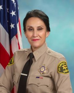 Commander Figueroa is wearing a tan long sleeve shirt with a black tie, badge on left side of shirt, sitting in front of a blue background. There is a hanging American flag in the background behind him on his right side.