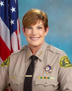 Assistant Sheriff Tokar is wearing a tan long sleeve shirt with a black tie, badge on left side of shirt, sitting in front of a blue background.