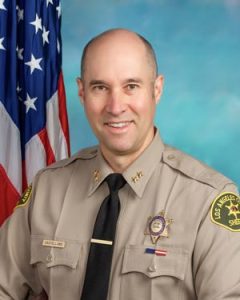 Chief Castellano is wearing a tan long sleeve shirt with a black tie, badge on left side of shirt, sitting in front of a blue background. There is a hanging American flag in the background behind him on his right side.