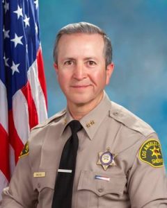 Chief Macias is wearing a tan long sleeve shirt with a black tie, badge on left side of shirt, sitting in front of a blue background. There is a hanging American flag in the background behind him on his right side.