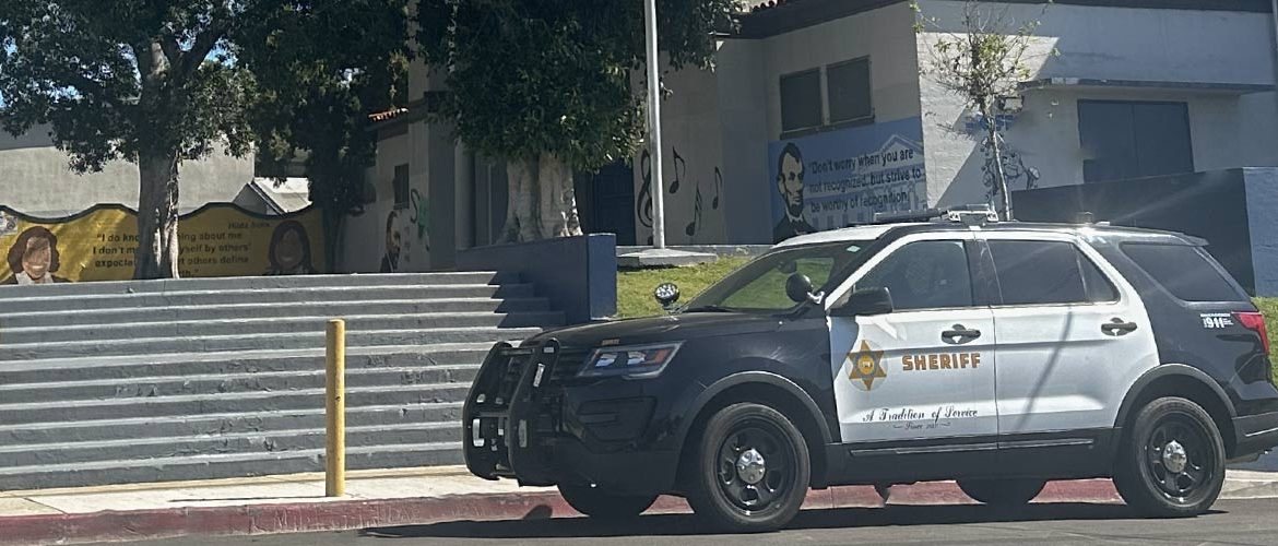 Sheriff's black and white truck with a sheriff's star logo on the door is parked on the curb in front of a school's entrance. Stairs are seen going up to the quad with murals painted on the walls of historic figures and quotes.