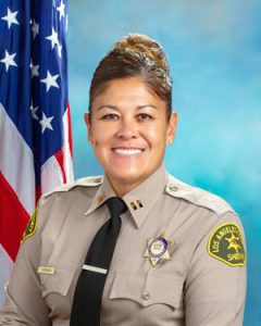 Captain Guerrero is wearing a tan long sleeve shirt with a black tie, badge on left side of shirt, sitting in front of a blue background, the american flag is behind him, over his right shoulder.