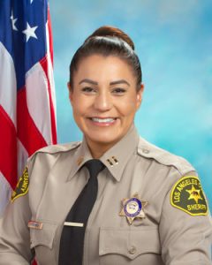 Captain Hinchman is wearing a tan long sleeve shirt with a black tie, badge on left side of shirt, sitting in front of a blue background, the american flag is behind him, over his right shoulder.