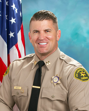 Captain Barclay is wearing a tan long sleeve shirt with a black tie, badge on left side of shirt, sitting in front of a blue background, the american flag is behind him, over his right shoulder.