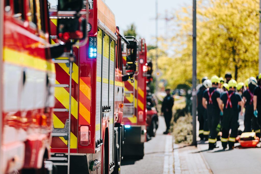 Several fire trucks are parked on a street in an urban setting, with a group of firefighters in helmets and dark uniforms standing nearby. The firetrucks are red with yellow and orange stripes. A person in dark clothing stands slightly apart from the group of firefighters. The lighting suggests it is daytime.
