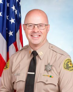 Captain Jones is wearing a tan long sleeve shirt with a black tie, badge on left side of shirt, sitting in front of a blue background. There is a hanging American flag in the background behind him on his right side.