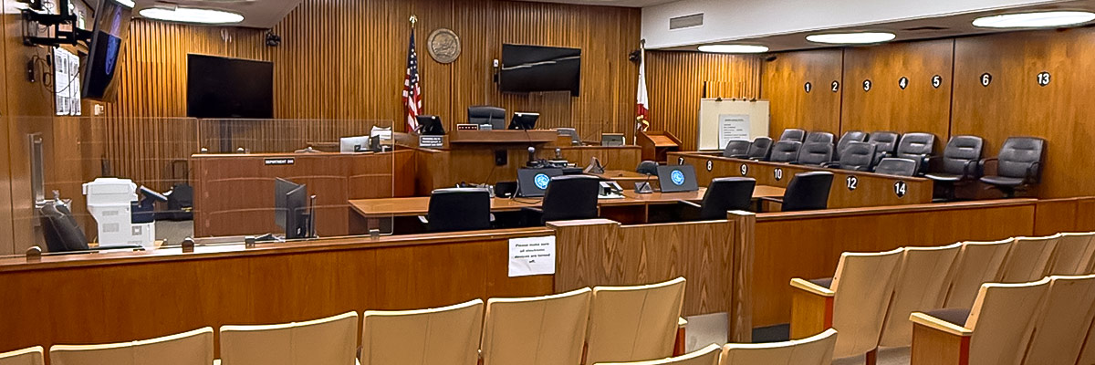 image of interior of a court room. wood paneled room with a partition in the front of the room separating single chairs in the gallery from the court. Two tables are on the other side of the partition. A raised partition has the judges chair and on either side are chairs for clerks and bailiffs.