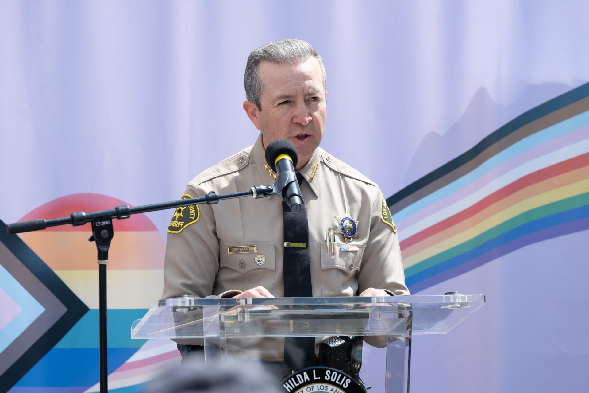 Assistant Sheriff Macias is speaking at a podium infront of a pride flag.