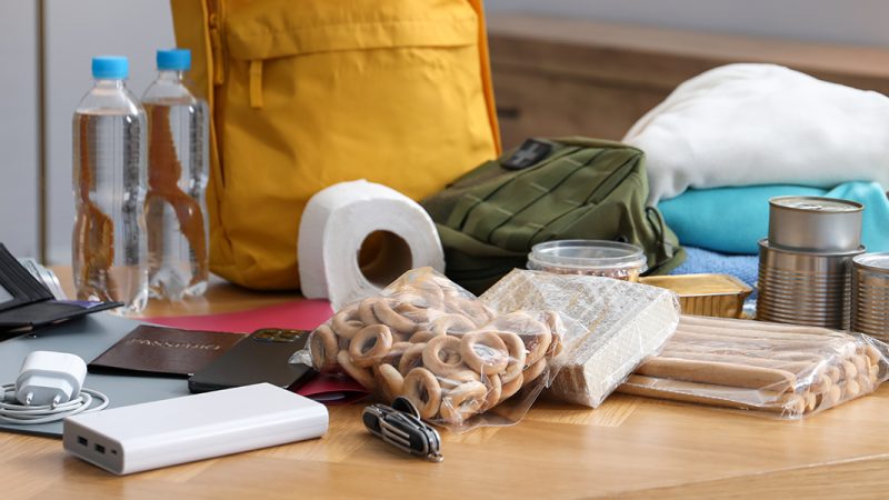 supplies and a yellow backpack displayed on a table. Charger, battery pack, bagged food and clothes.